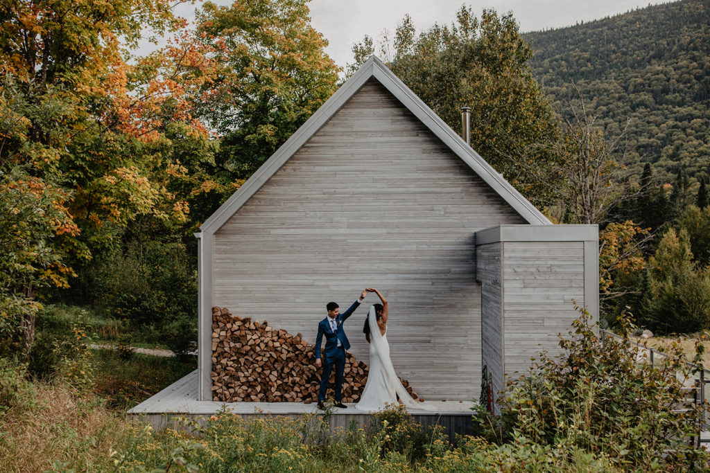 Couple twirling in wedding attire, in front of a wood pile and cabin